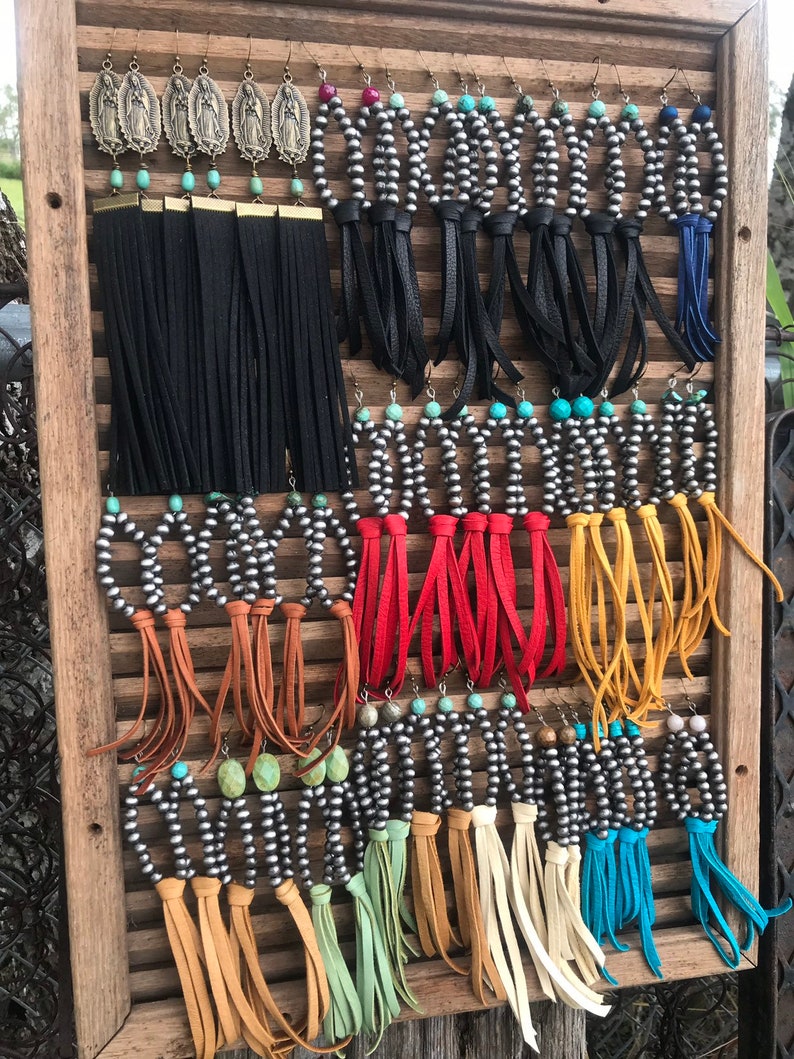 May include: A display of silver and turquoise earrings with leather fringe. The earrings are arranged on a wooden board with a slatted design. The fringe is in a variety of colors, including black, red, yellow, brown, and blue.