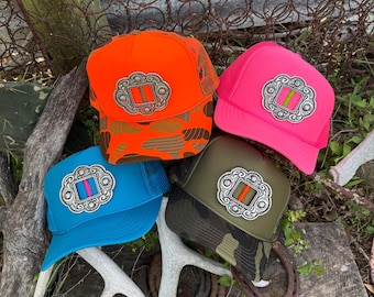 Puede incluir: Cuatro gorras tipo trucker con emblemas plateados estilo concho en los paneles frontales. Las gorras son de color naranja neón con camuflaje marrón, rosa neón, azul brillante y verde oliva con patrones de camuflaje, cada una con un acento vertical.