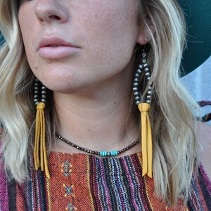 May include: A woman wearing a colorful striped top with a yellow leather tassel necklace and earrings. The earrings are made of silver beads and have a turquoise stone accent.