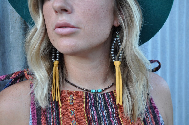 May include: A woman wearing a colorful striped top with a yellow leather tassel necklace and earrings. The earrings are made of silver beads and have a turquoise stone accent.
