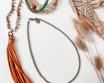 Puede incluir: Un collar de cuentas plateadas con un borla de cuero marrón cuelga junto a una pila de pulseras con cuentas verdes, beige y blancas. Un pañuelo marrón con un patrón blanco está en el fondo.