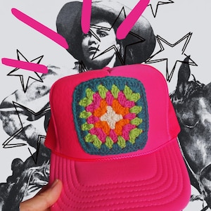 May include: A bright pink trucker hat with a crocheted square patch featuring a colorful floral design. The hat has a mesh back and a curved brim. The background includes a black and white image of a person wearing a hat and star illustrations.
