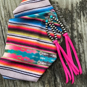 May include: A pair of silver tone earrings with turquoise accents and hot pink tassels. The earrings are displayed on a colorful striped fabric.