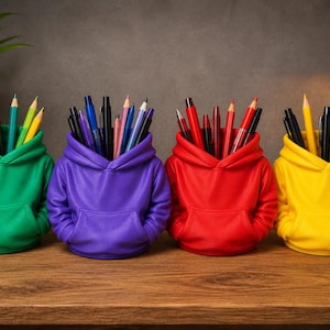 May include: Four desk organizers shaped like miniature hoodies in green, purple, red, and yellow. Each container features a hood, long sleeves, and a front kangaroo pocket, and is filled with various colored pens, pencils, and markers for storage.
