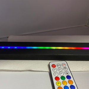 May include: A black LED light bar with a spectrum of colors, including blue, green, yellow, and red. A remote control with multiple buttons is in the foreground. The light bar is on a white surface.