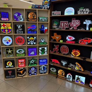 May include: A display of illuminated signs featuring various logos and designs. The signs include sports team emblems, Halloween themes, and phrases like "Bar Open" and "Star Wars." The signs are arranged on wooden shelves.