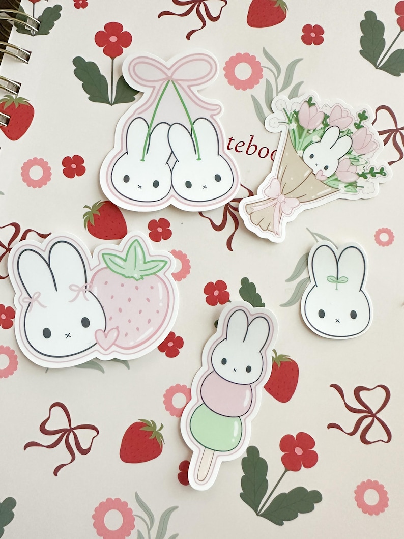 May include: A set of five stickers featuring cute bunny illustrations. The stickers are on a pink and white background with a floral pattern. The stickers include a bunny holding a strawberry, a bunny on a stick with green and pink mochi, two bunnies hanging from a pink ribbon, a bunny in a bouquet of flowers, and a single bunny.