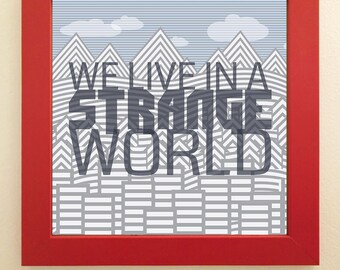 May include: A framed art print with a red frame. The print features a mountain landscape design in shades of gray and white, with the text "WE LIVE IN A STRANGE WORLD" in a bold, stylized font. The background is a light blue with white clouds.