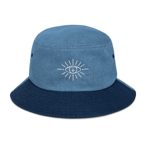 Op de afbeelding: Een blauwe denim bucket hat met een wit geborduurd oogontwerp. De hoed heeft een donkerblauwe rand.