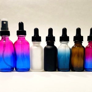May include: A collection of glass bottles with black droppers and spray tops. The bottles are in various colors, including pink and blue gradients, white, black, blue, and amber. The bottles are arranged in a row against a white background.
