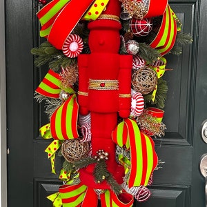 May include: A festive Christmas door decoration featuring a large red nutcracker figure. The decoration includes red and green striped ribbons, gold accents, and decorative ornaments. The door is painted a dark green.