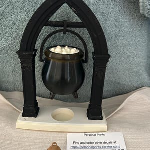May include: A black cauldron with a white bubbling liquid inside, suspended from a black gothic-style arch frame on a white base. A small tan figurine sits in front of a white card with the text: Personal Prints Find and order other decals at: https://personalprints.ecrater.com/ SCAN HERE: and a QR code.
