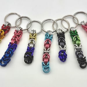 May include: A collection of seven keychains with colorful, chainmail-style pendants. The pendants feature a variety of colors, including rainbow, pink, blue, black, and green, attached to silver key rings. The keychains are displayed against a white background.