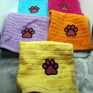 May include: A selection of colourful, square hand towels. Each towel is a different colour: blue, pink, orange, purple, and yellow. Each towel has an embroidered paw print in red with a black outline.