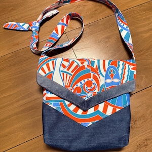 May include: A crossbody bag featuring a dark blue denim lower section and a flap with a vibrant geometric pattern in orange, light blue, and white. The bag includes a matching patterned shoulder strap with a knot detail at the top for adjustment.