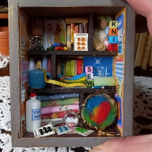Puede incluir: Miniatura de habitación artesanal de madera gris con estantes llenos de suministros de costura y artesanía. Incluye carretes de hilo, retales de tela, botones, una taza azul y una botella de pegamento PVA. La palabra "SEW" es visible.