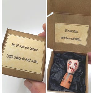 May include: A small brown cardboard box containing a miniature figurine nestled in black tissue paper. The lid exterior reads, "We all have our demons, I just choose to feed mine.." and the interior lid reads, "This one likes milkshake and chips."