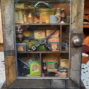 Könnte beinhalten: Ein Miniatur-Holzschrank mit Glasfront, gefüllt mit detaillierten Miniaturartikeln. Der Schrank hat ein rustikales, gealtertes Aussehen. Zu den Artikeln gehören Miniatur-Lebensmittelverpackungen, Flaschen und eine Spielzeugpistole. Der Text "NOT FOOD" ist auf einer Flasche zu sehen.