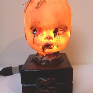 May include: A spooky doll head lamp with a glowing orange light. The doll's face has dark streaks and appears to be crying. It sits atop a black box with a metal clasp, and a USB cable extends from the base.