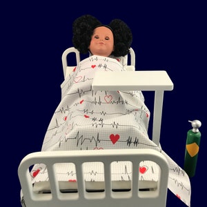 May include: A white doll bed with a doll lying in it. The doll has black hair and is wearing a white and red sheet with a heart beat pattern. There is a white table next to the bed and a green oxygen tank on the floor.