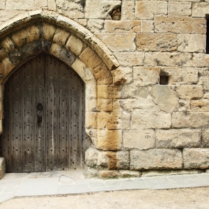 May include: A weathered wooden door set within a stone archway. The door is made of vertical wooden planks with metal hardware. The archway and surrounding wall are constructed of light brown stone blocks, with a small window visible.