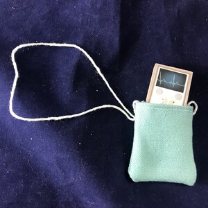 May include: A small, white, electronic device with a heart rate monitor display and two buttons. The device is in a light blue drawstring bag with a white cord.