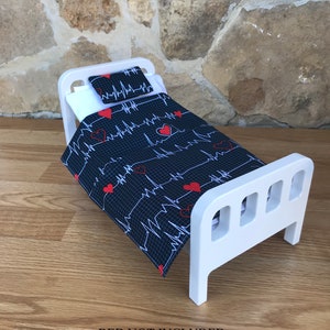 May include: A white doll bed with a black and white bedding set featuring red hearts and EKG lines. The bedding set includes a pillow and a comforter. "BED NOT INCLUDED" is written in black text on the wooden floor.