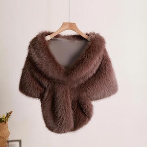 Peut inclure: Un châle en fausse fourrure marron avec une texture douce et moelleuse. Le châle est drapé sur un cintre en bois et présente un design simple et élégant.