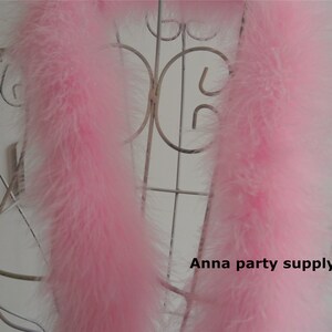 May include: A long, pink feather boa draped over a white metal display stand. The boa is soft and fluffy, and it is perfect for adding a touch of glamour to any outfit or event.  Anna party supply.