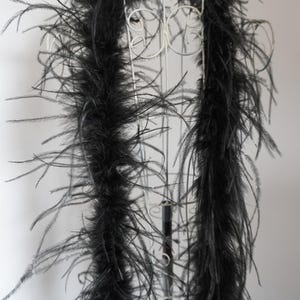 May include: Black feather boa draped over a white metal display stand. The boa is thick and fluffy, with long, soft feathers. The stand is ornate, with a circular top and decorative swirls. The overall effect is elegant and dramatic.