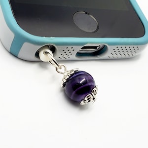Puede incluir: Un charm para conector de teléfono con una cuenta de ágata púrpura y detalles plateados. El charm está unido a un teléfono inteligente.