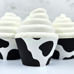 Può includere: Tre cupcake bianchi con pirottini da cupcake a stampa di mucca in bianco e nero. I cupcake sono glassati con glassa bianca.