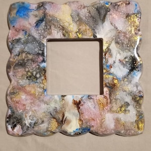 Puede incluir: Un marco de fotos blanco con un borde ondulado y festoneado. El marco está decorado con un diseño abstracto en espiral en tonos de rosa, dorado, marrón y azul. El diseño incluye purpurina y pequeñas burbujas de aire.
