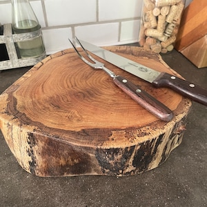 May include: A large, round wooden cutting board made from a tree trunk slice. The board has a natural, rustic finish with dark brown bark around the edges. A carving knife and fork are placed on the board.