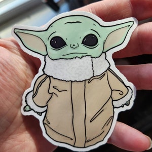 Puede incluir: Una pegatina verde y marr&oacute;n de un personaje de la franquicia Star Wars, conocido como "The Child" o "Baby Yoda", que lleva una chaqueta marr&oacute;n con un cuello de piel blanca.