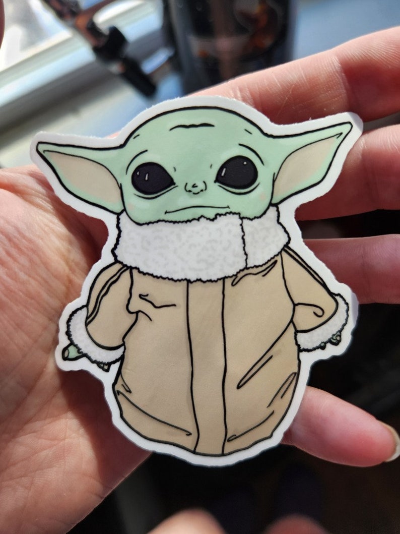 Puede incluir: Una pegatina verde y marr&oacute;n de un personaje de la franquicia Star Wars, conocido como "The Child" o "Baby Yoda", que lleva una chaqueta marr&oacute;n con un cuello de piel blanca.