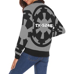 May include: Gray bomber jacket featuring a large black and gray Imperial symbol on the back. The jacket has black cuffs, a black hem, and the text "TK-5246" in white. The jacket is worn over blue jeans.
