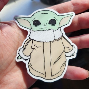 Puede incluir: Una pegatina verde y beige de un personaje de la franquicia Star Wars, conocido como "The Child" o "Baby Yoda", que lleva una chaqueta marr&oacute;n con un cuello de piel blanca. El personaje tiene ojos grandes y oscuros y una expresi&oacute;n seria.
