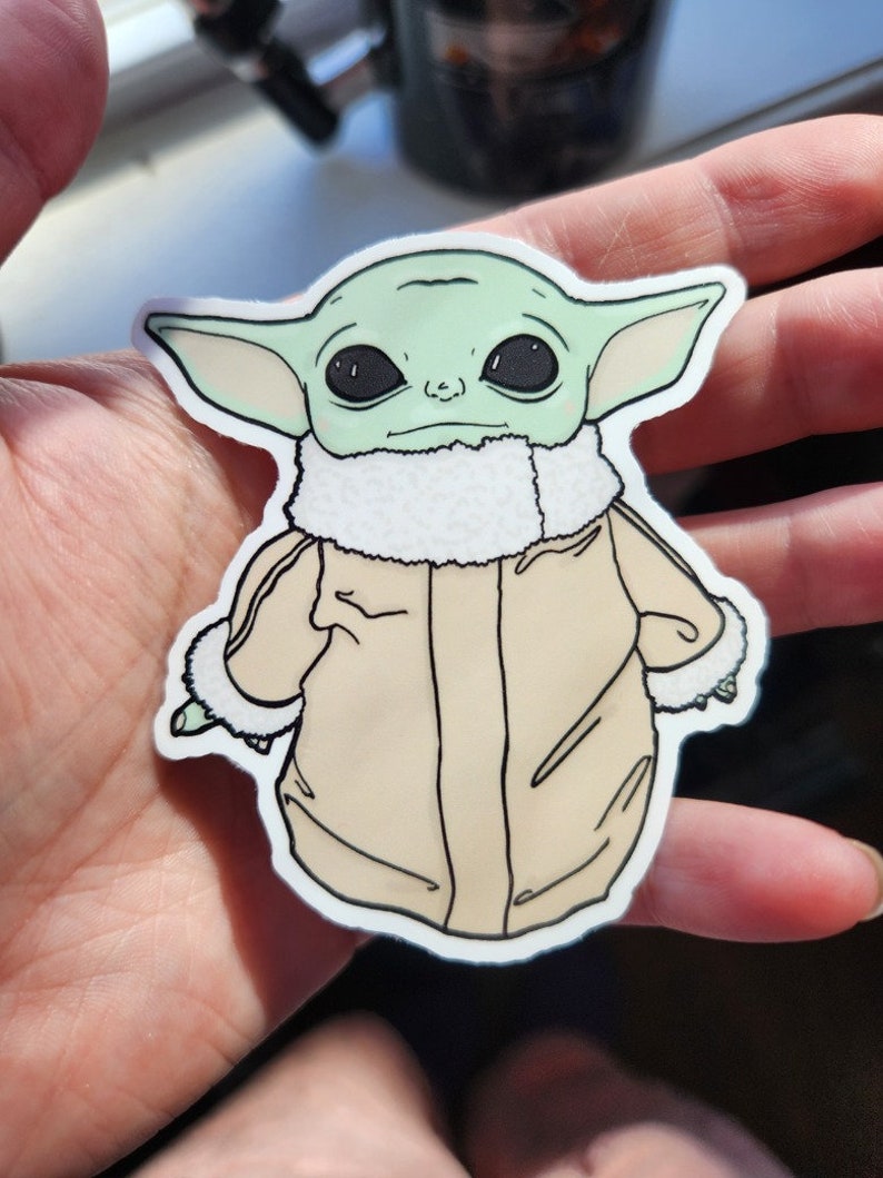 Puede incluir: Una pegatina verde y beige de un personaje de la franquicia Star Wars, conocido como "The Child" o "Baby Yoda", que lleva una chaqueta marr&oacute;n con un cuello de piel blanca. El personaje tiene ojos grandes y oscuros y una expresi&oacute;n seria.
