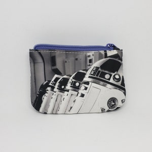 Könnte beinhalten: Eine schwarze und weiße Reißverschlusstasche mit einem sich wiederholenden Bild von Star Wars R2-D2 Robotern. Die Tasche hat einen lila Reißverschluss.