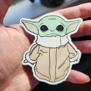Puede incluir: Una pegatina verde y beige de un personaje de dibujos animados con orejas grandes, que lleva una chaqueta marr&oacute;n con un cuello de piel blanca. El personaje tiene ojos negros grandes y una boca peque&ntilde;a.