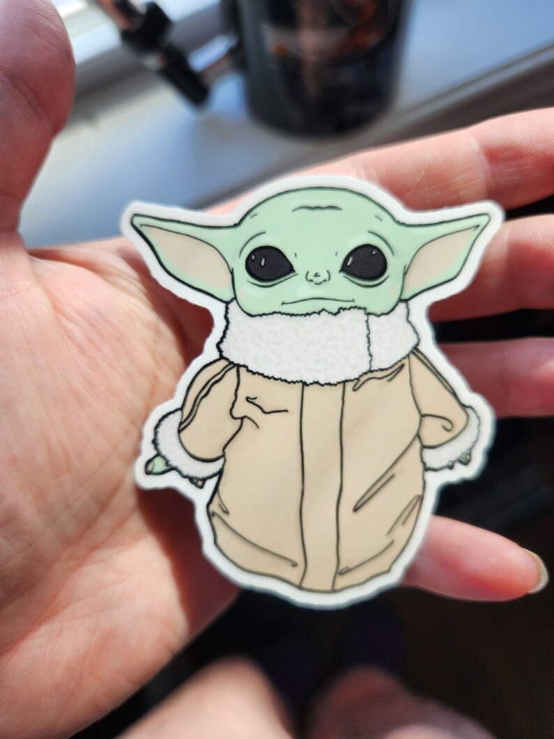 Puede incluir: Una pegatina verde y beige de un personaje de dibujos animados con orejas grandes, que lleva una chaqueta marr&oacute;n con un cuello de piel blanca. El personaje tiene ojos negros grandes y una boca peque&ntilde;a.