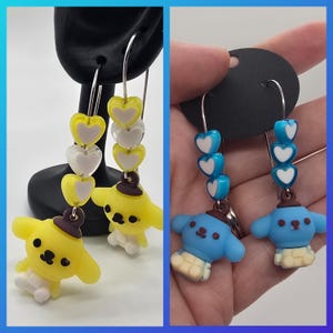 Op de afbeelding: Dangle oorbellen met cartoonhond bedels. Eén paar is geel met een bruine hoed, de andere is blauw met een bruine hoed. Beide paren hebben hartvormige kralen in geel, wit en helder of blauw en wit.
