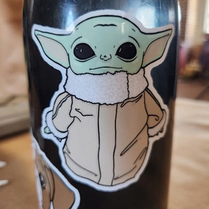 Puede incluir: Una botella de agua negra con una pegatina verde y marr&oacute;n de un personaje de la franquicia Star Wars. El personaje lleva una chaqueta marr&oacute;n con un cuello de piel blanca.