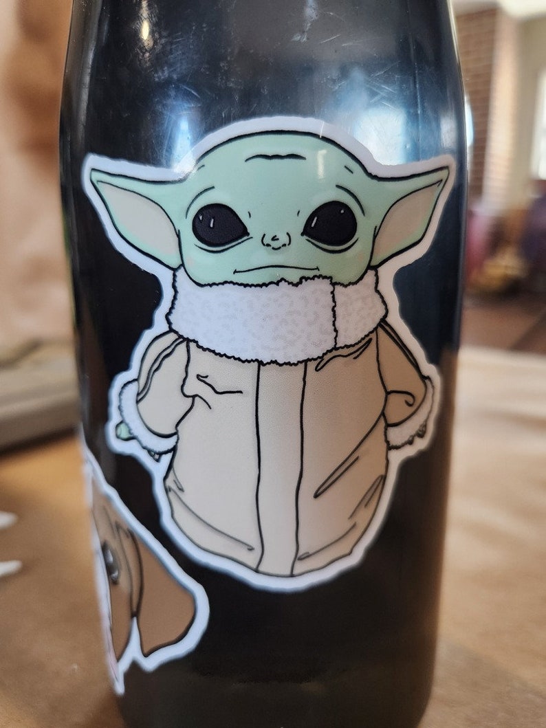 Puede incluir: Una botella de agua negra con una pegatina verde y marr&oacute;n de un personaje de la franquicia Star Wars. El personaje lleva una chaqueta marr&oacute;n con un cuello de piel blanca.