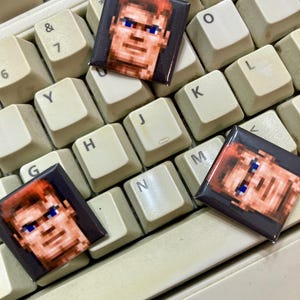 May include: Three pixelated character portraits on square pins, each with a red-brown hair, blue eyes, and a pixelated face. The pins are placed on a white keyboard.