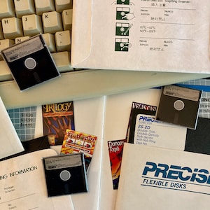 May include: A close-up of a stack of floppy disks, including a black and white floppy disk with the text "The Oregon Trail" and a black floppy disk with the text "Vegas Casino". The floppy disks are on a white surface with a keyboard and a brown envelope in the background.