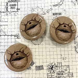 May include: Three brown round buttons with a black and white illustration of a one-eyed monster with sharp teeth and tentacles. The buttons are on a white grid background with black lines.