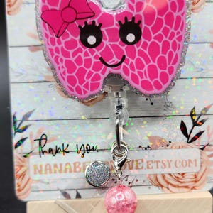 May include: Pink glitter thyroid shaped badge reel with a bow and a smiling face. The reel is attached to a silver charm with a pink and clear crystal bead. The text "Nana Bear Love" is visible in the background.