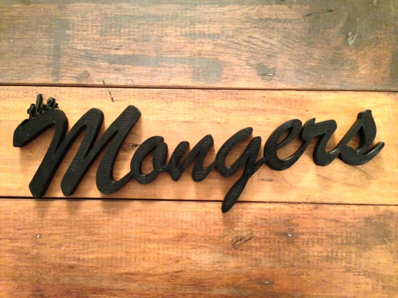 K&ouml;nnte beinhalten: Schwarzes Holzschild mit dem Wort "Mongers" in Schreibschrift. Das Schild liegt auf einer Holzoberfl&auml;che mit nat&uuml;rlicher Maserung.